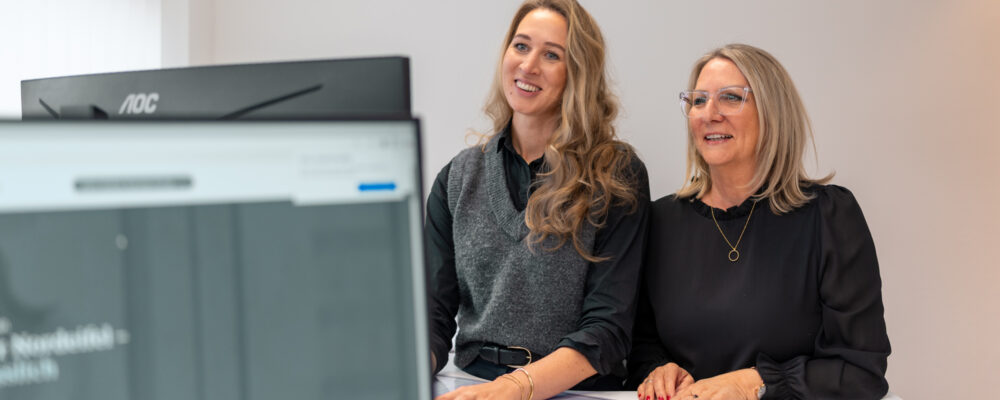 Zwie Frauen sitzen am Schreibtisch vor einem Monitor und tauschen sihc aus.