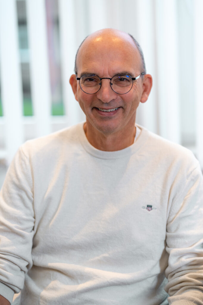 Mann mit Brille und weissem Pullover sitzt und laechelt in die Kamera
