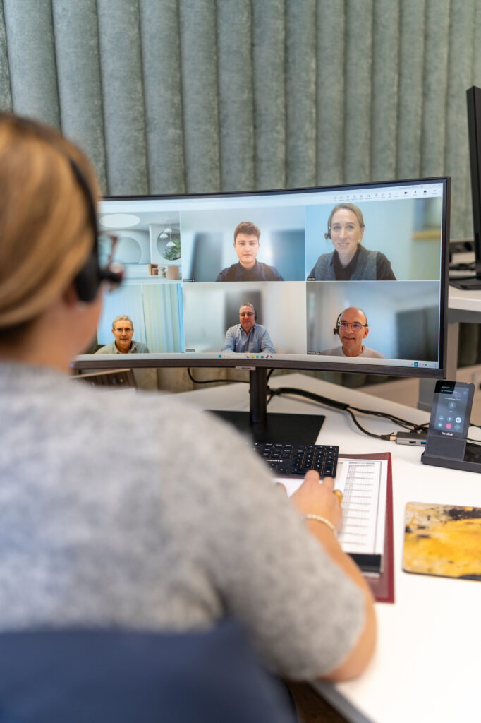 Person mit Headset sitzt am Schreibtisch vor einem grossen Monitor mit einer Videokonferenz